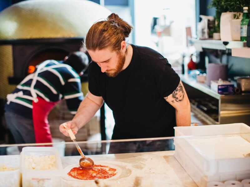 A man in a black t-shirt adding sauce to a pizza base